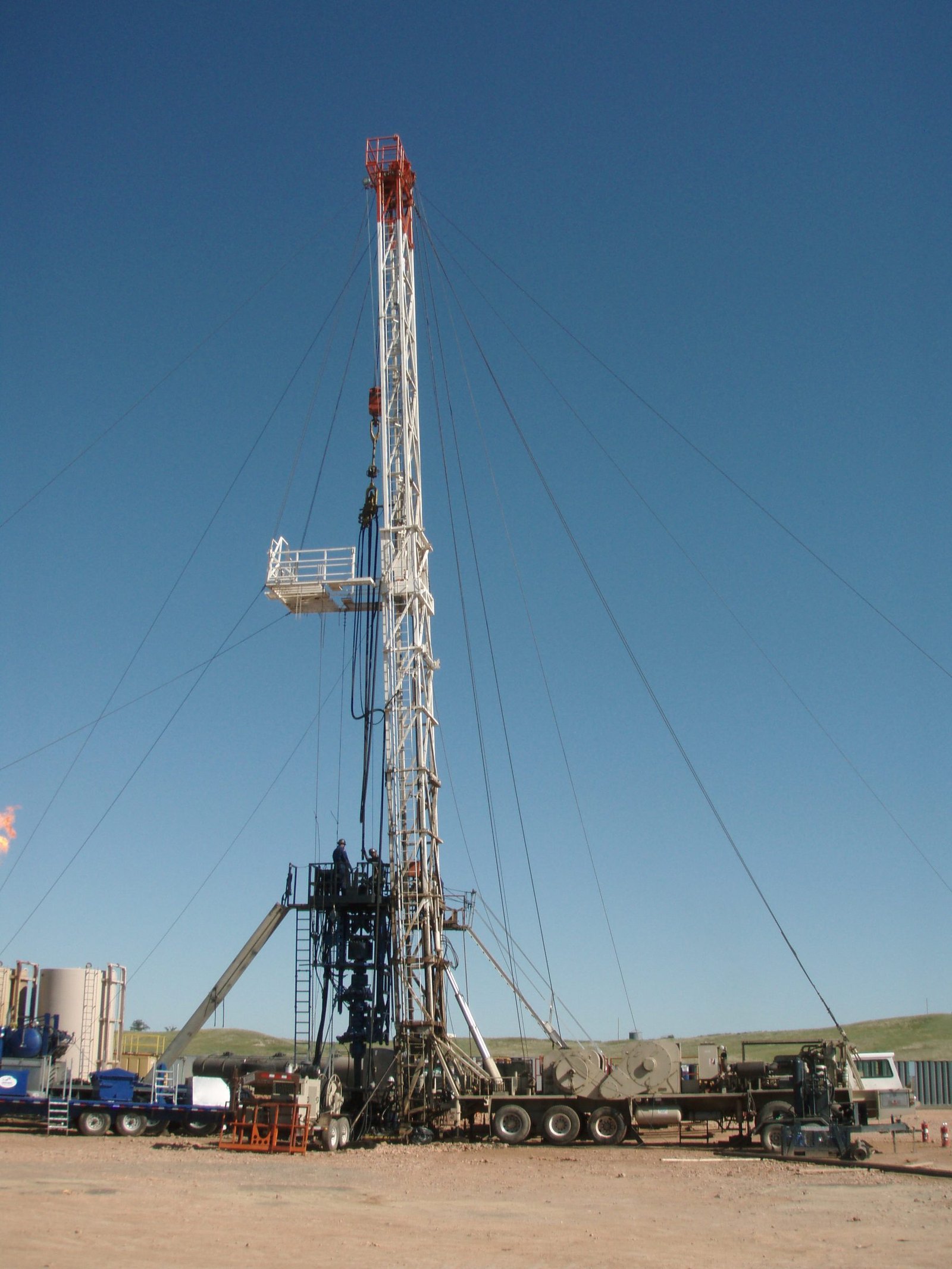 A tall service rig standing over a wellhead on a gravel location under a blue sky